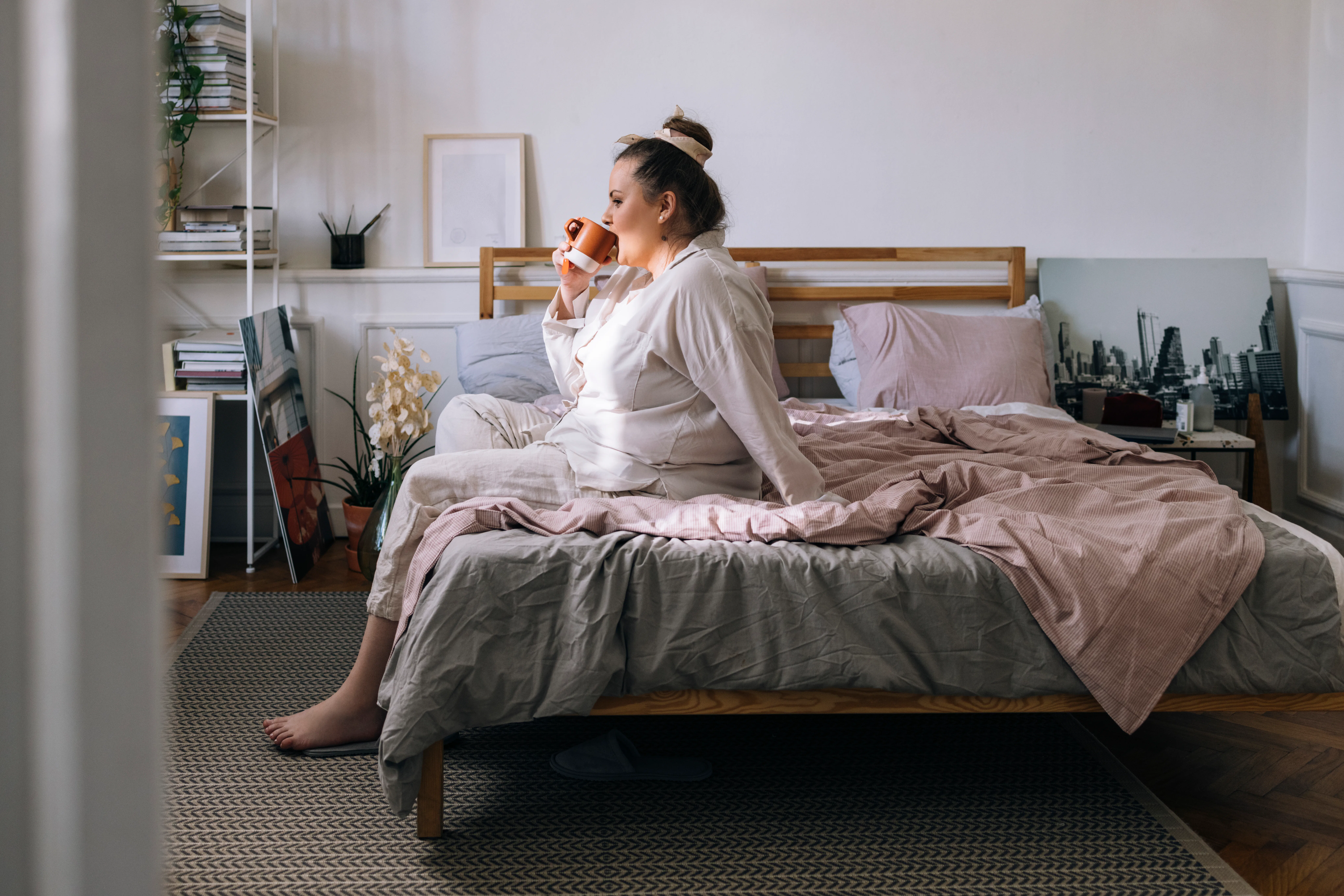 A young Caucasian plus size woman sitting on the bed in her pyjamas and having a cup of coffee in th...