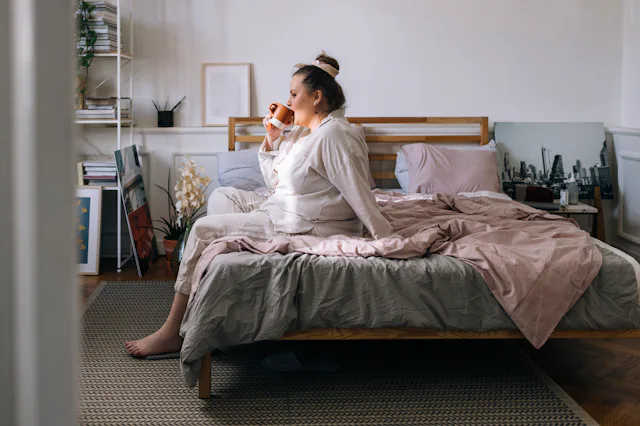 A young Caucasian plus size woman sitting on the bed in her pyjamas and having a cup of coffee in th...