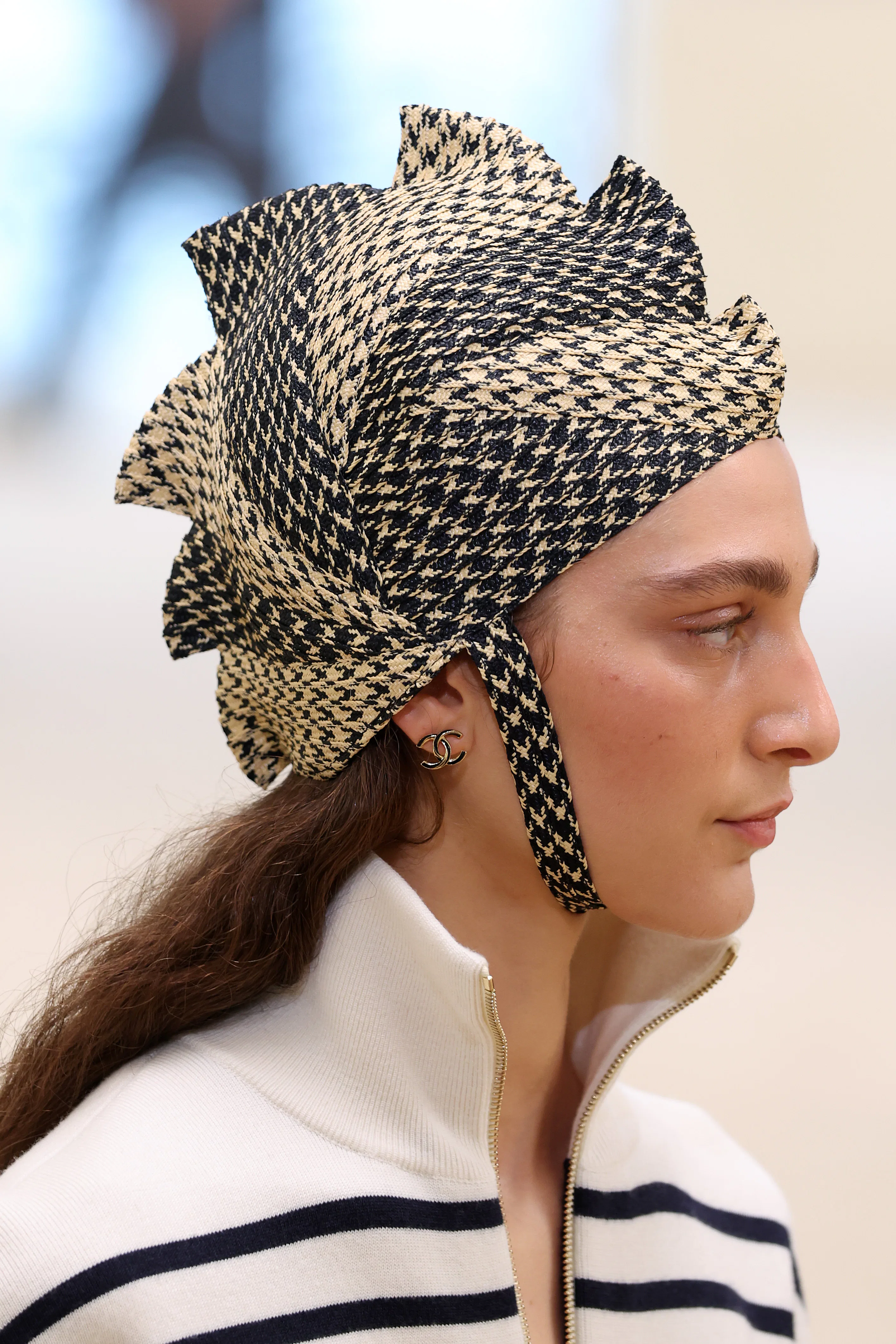 Şapkasının detaylarını sergileyen bir manken, Chanel Cruise 2026/27 defilesinde podyumda yürüyor.