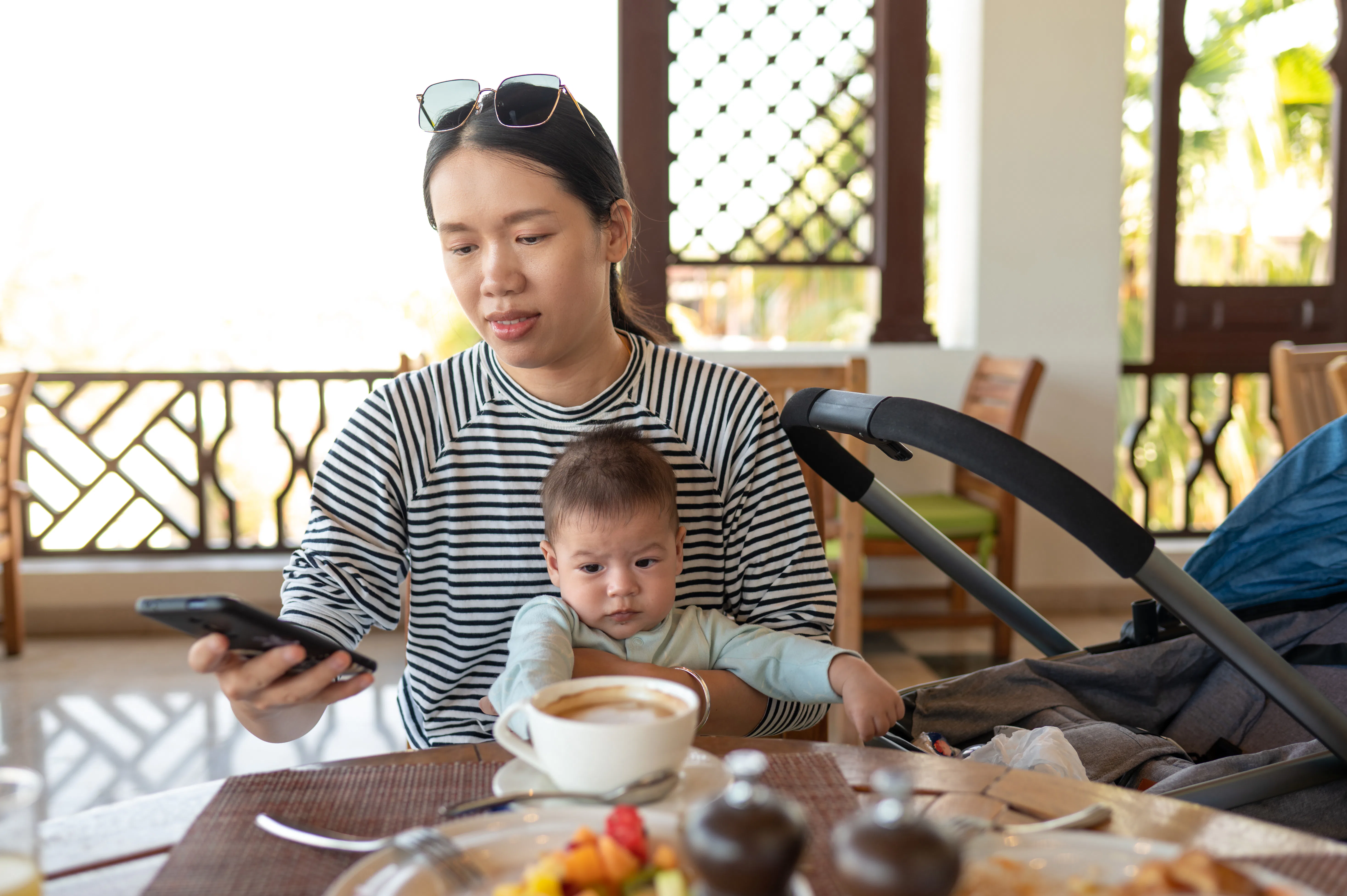 Young Asian woman using phone while having a cup coffee in a restaurant holding with her newborn bab...