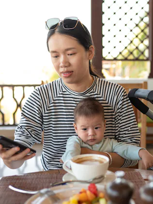 Young Asian woman using phone while having a cup coffee in a restaurant holding with her newborn bab...