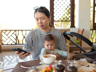 Young Asian woman using phone while having a cup coffee in a restaurant holding with her newborn bab...