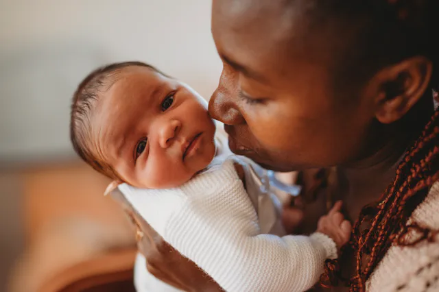 A person tenderly holds and kisses a baby on the cheek indoors. The baby, wearing a white outfit, ga...