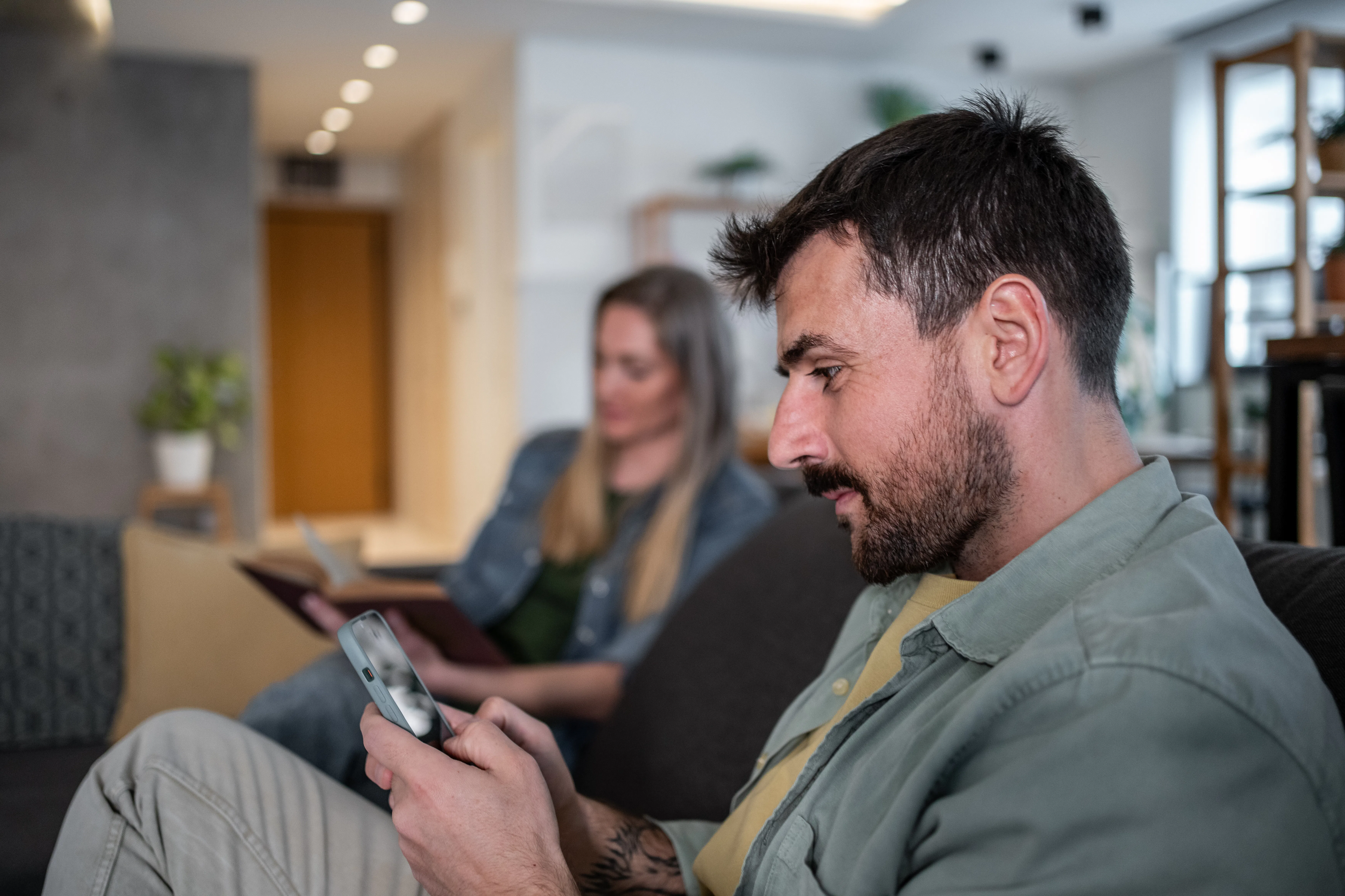 Man using smartphone while woman reading book, sitting together on sofa in living room, ignoring eac...