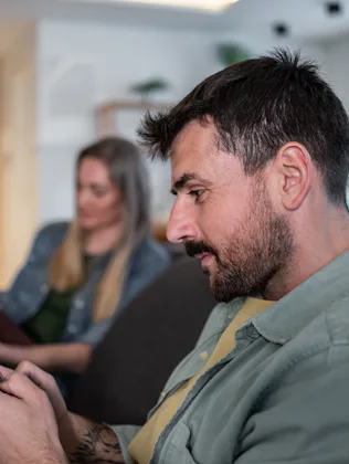 Man using smartphone while woman reading book, sitting together on sofa in living room, ignoring eac...