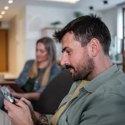 Man using smartphone while woman reading book, sitting together on sofa in living room, ignoring each other, modern lifestyle and technology addiction concept