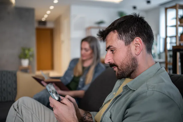 Man using smartphone while woman reading book, sitting together on sofa in living room, ignoring eac...