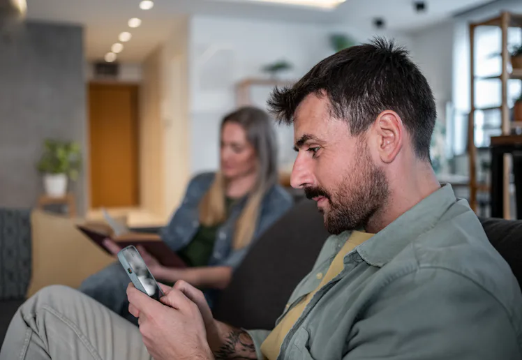 Man using smartphone while woman reading book, sitting together on sofa in living room, ignoring eac...