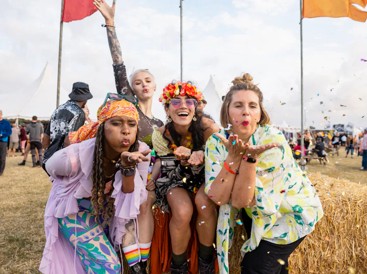 Group photo of four female friends blowing confetti into the camera lens. They are all dressed in vi...