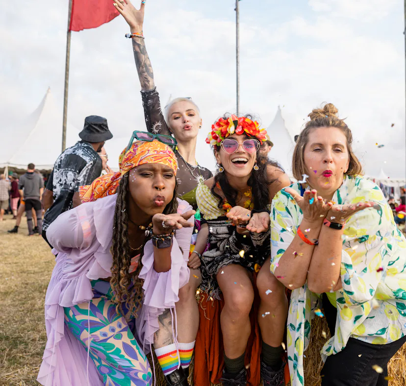 Group photo of four female friends blowing confetti into the camera lens. They are all dressed in vi...