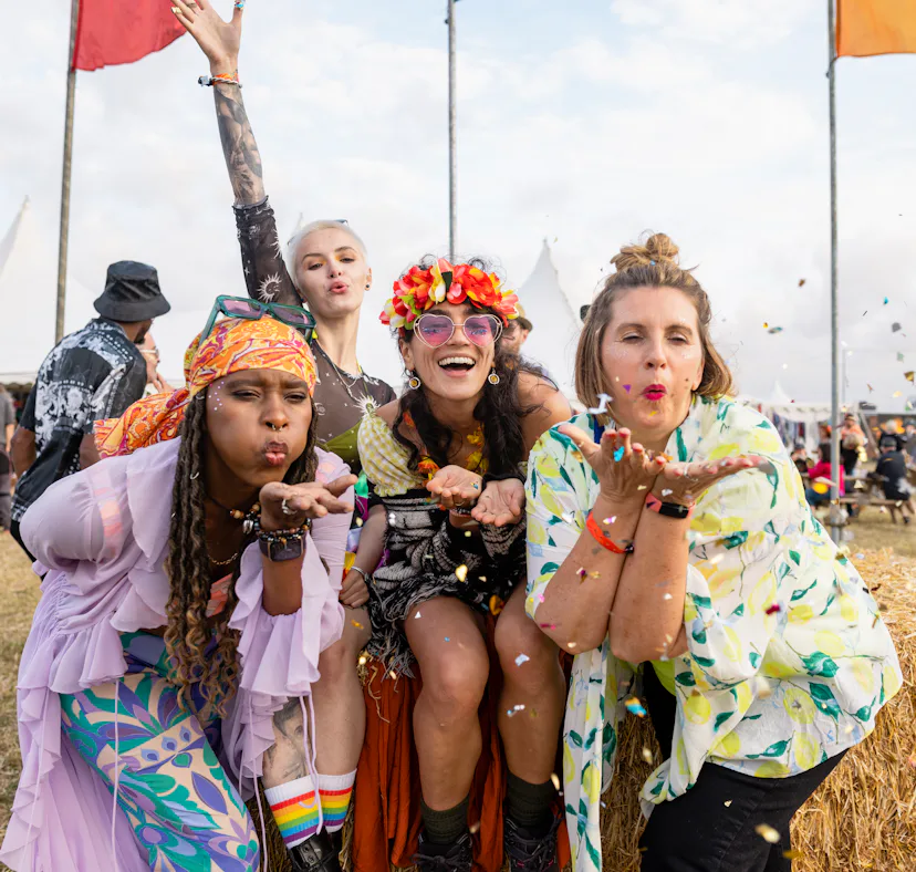 Group photo of four female friends blowing confetti into the camera lens. They are all dressed in vi...