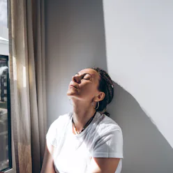 Portrait of a dreadlock braid woman sitting by the open window at the white wall. Concept of depress...