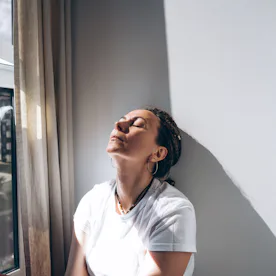 Portrait of a dreadlock braid woman sitting by the open window at the white wall. Concept of depress...