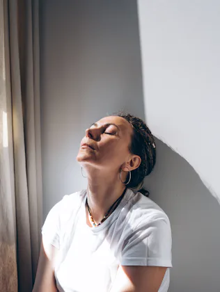 Portrait of a dreadlock braid woman sitting by the open window at the white wall. Concept of depress...