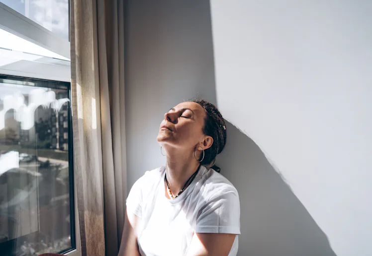Portrait of a dreadlock braid woman sitting by the open window at the white wall. Concept of depress...