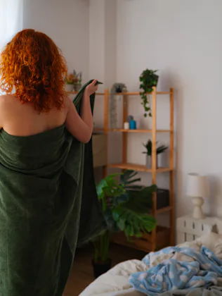 Young redhead stands confidently in bedroom, wrapping towel around body after shower. She embraces m...