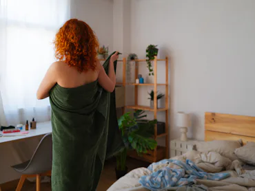 Young redhead stands confidently in bedroom, wrapping towel around body after shower. She embraces m...