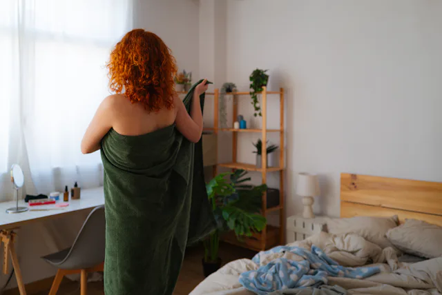 Young redhead stands confidently in bedroom, wrapping towel around body after shower. She embraces m...