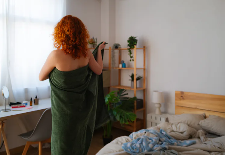 Young redhead stands confidently in bedroom, wrapping towel around body after shower. She embraces m...