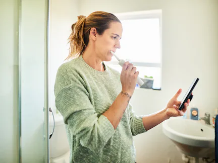Woman standing in her bathroom at home brushing her teeth with an electric toothbrush and checking h...