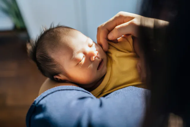 Over the shoulder view of Asian mother craddles her sleeping baby in her arms and gently touching he...
