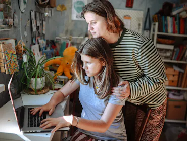 Plus-size middle-aged mom and teenage daughter working together on a laptop at home, academic suppor...