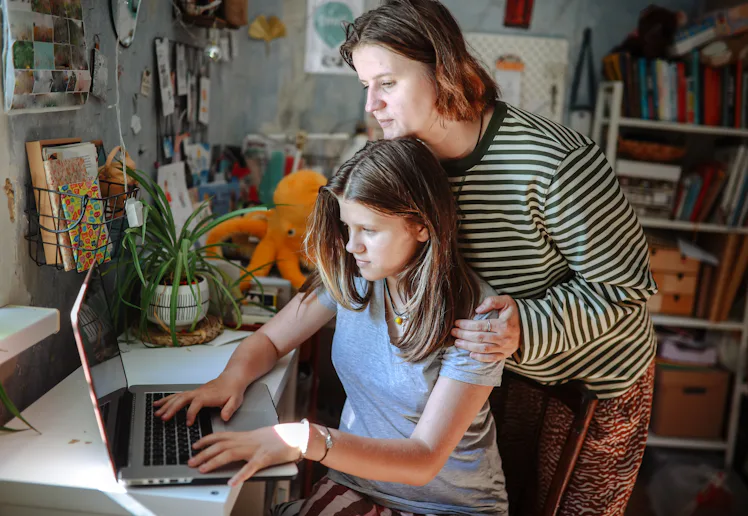 Plus-size middle-aged mom and teenage daughter working together on a laptop at home, academic suppor...