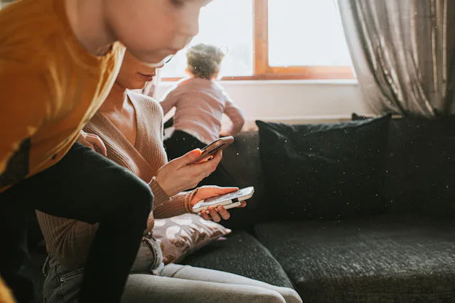 A single mum sits on a sofa looking down at two mobile phones. She attempts to stay focused and work...