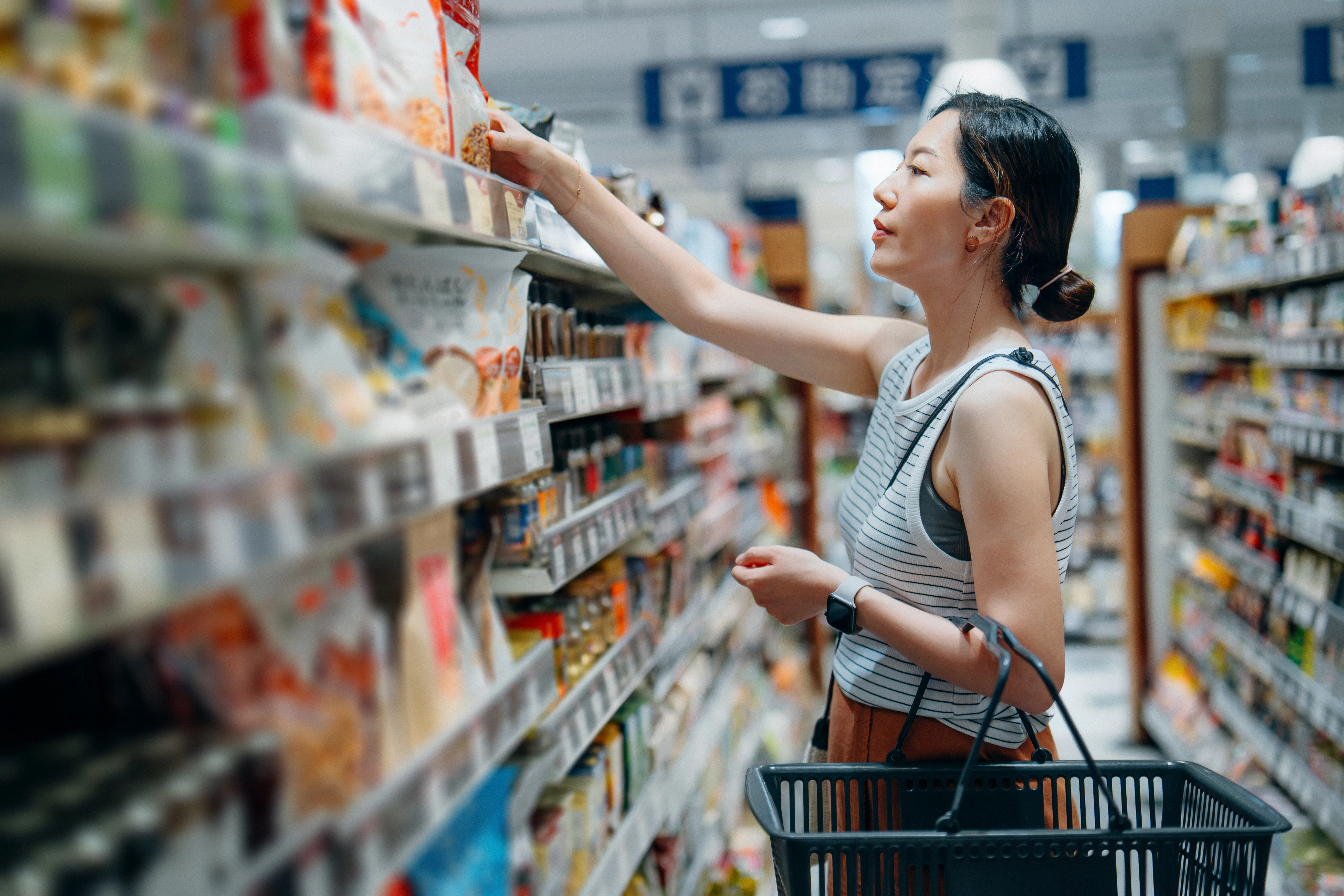Young Asian woman carrying a shopping basket, grocery shopping in supermarket. She is looking at pro...