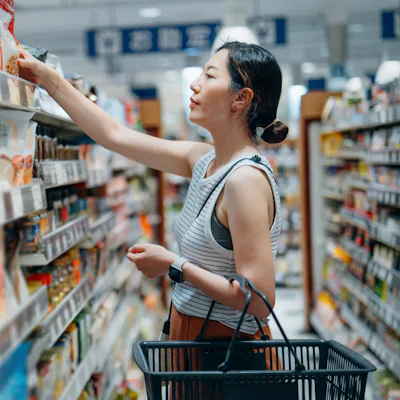 Young Asian woman carrying a shopping basket, grocery shopping in supermarket. She is looking at products on the shelf, choosing what she should buy. Routine grocery shopping. Making better and healthier food choices. Consumer awareness concept
