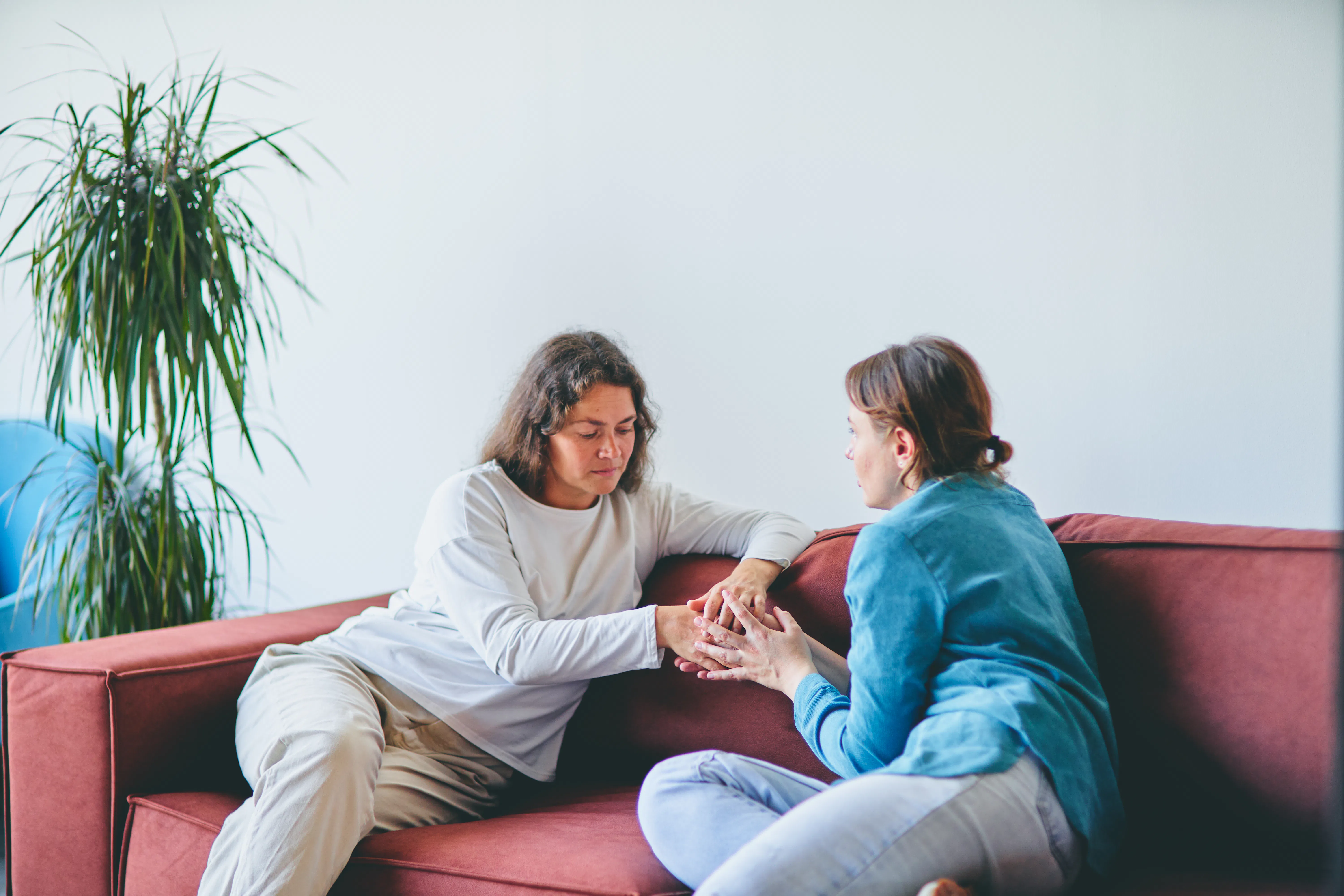In a modern living room, two women sit on a red sofa. The woman in the white shirt shows support and...