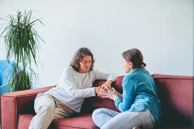 In a modern living room, two women sit on a red sofa. The woman in the white shirt shows support and...