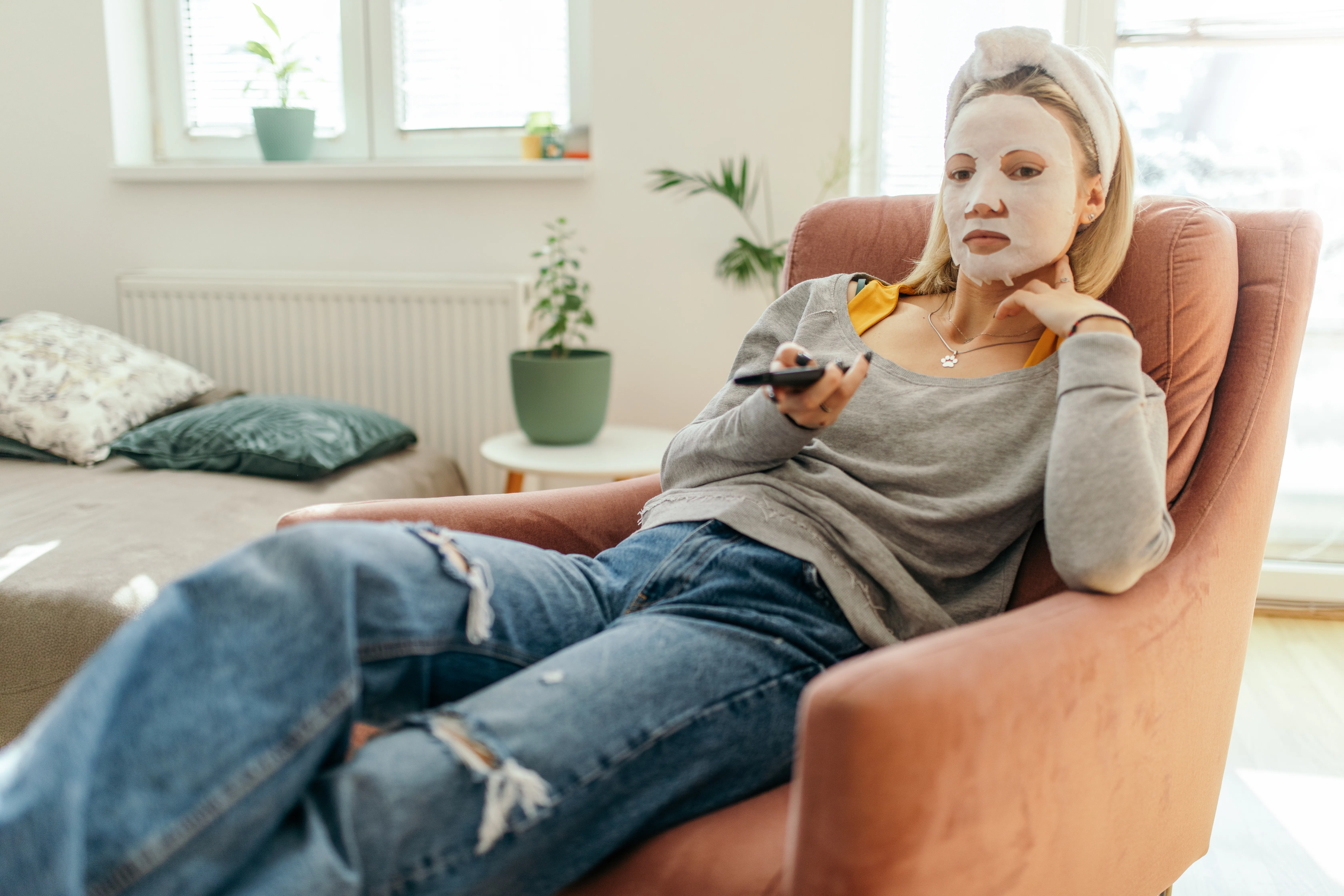 Young woman is sitting cozy on armchair at home while watching TV. She has a facial cosmetics mask o...