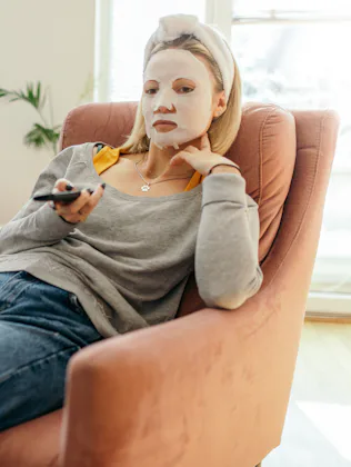 Young woman is sitting cozy on armchair at home while watching TV. She has a facial cosmetics mask o...