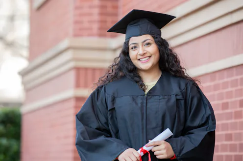 Happy Indian woman wearing graduation cap and gown, celebrating college success outdoors.