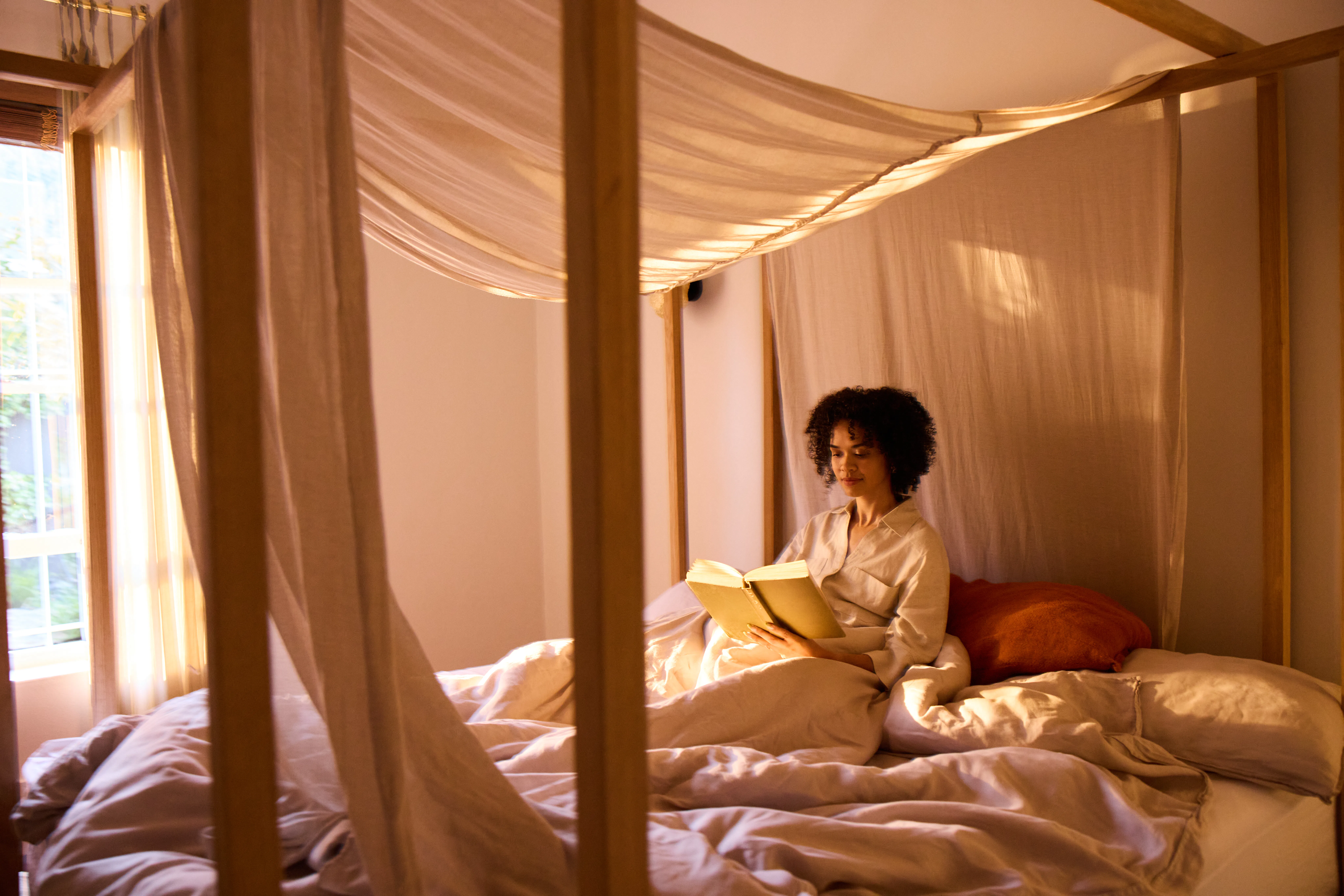 Wide shot of a young woman sitting on a canopy bed and reading a book quietly in a softly lit bedroo...