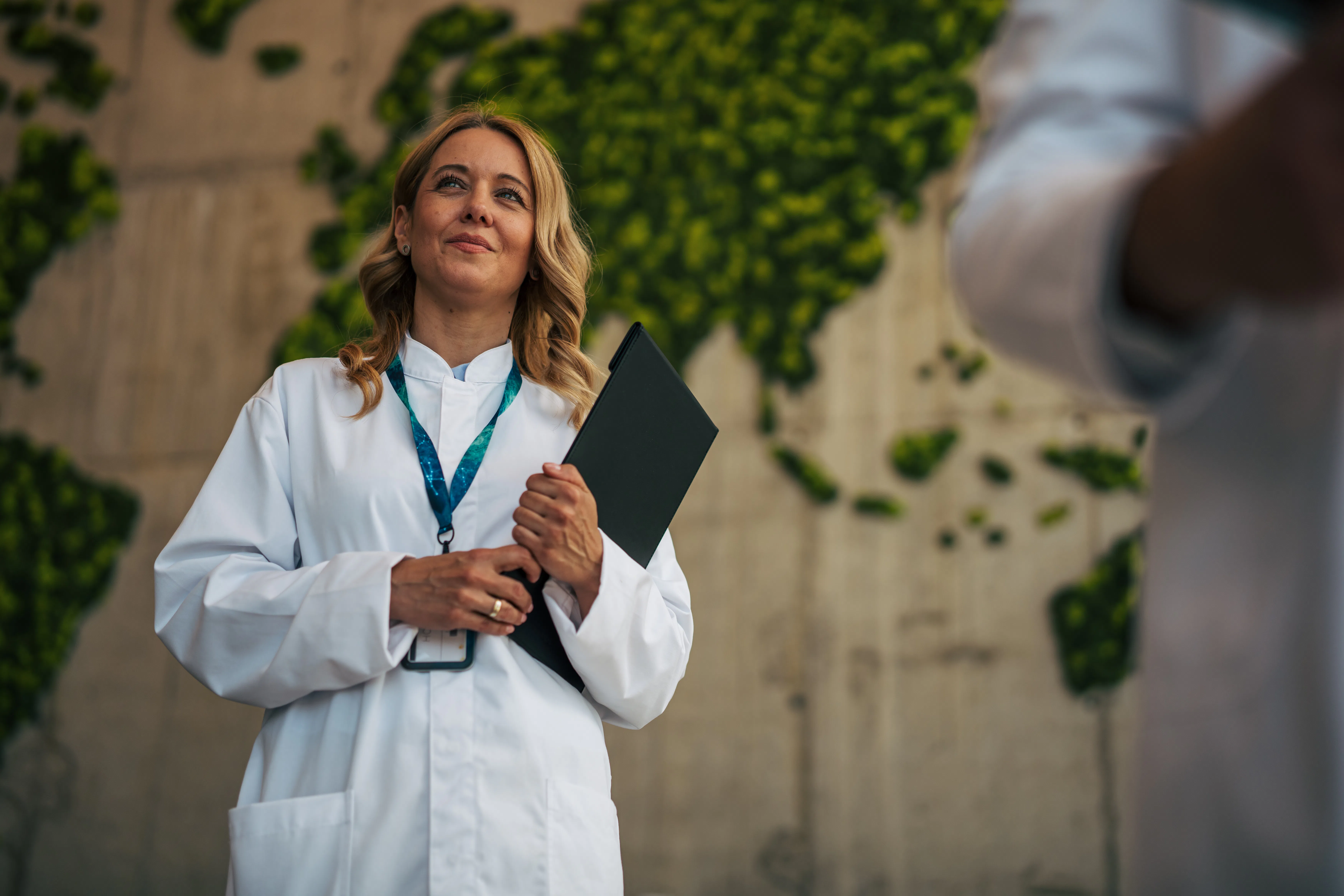 Female scientist holding a folder and looking up, standing in a modern office with a green living wo...