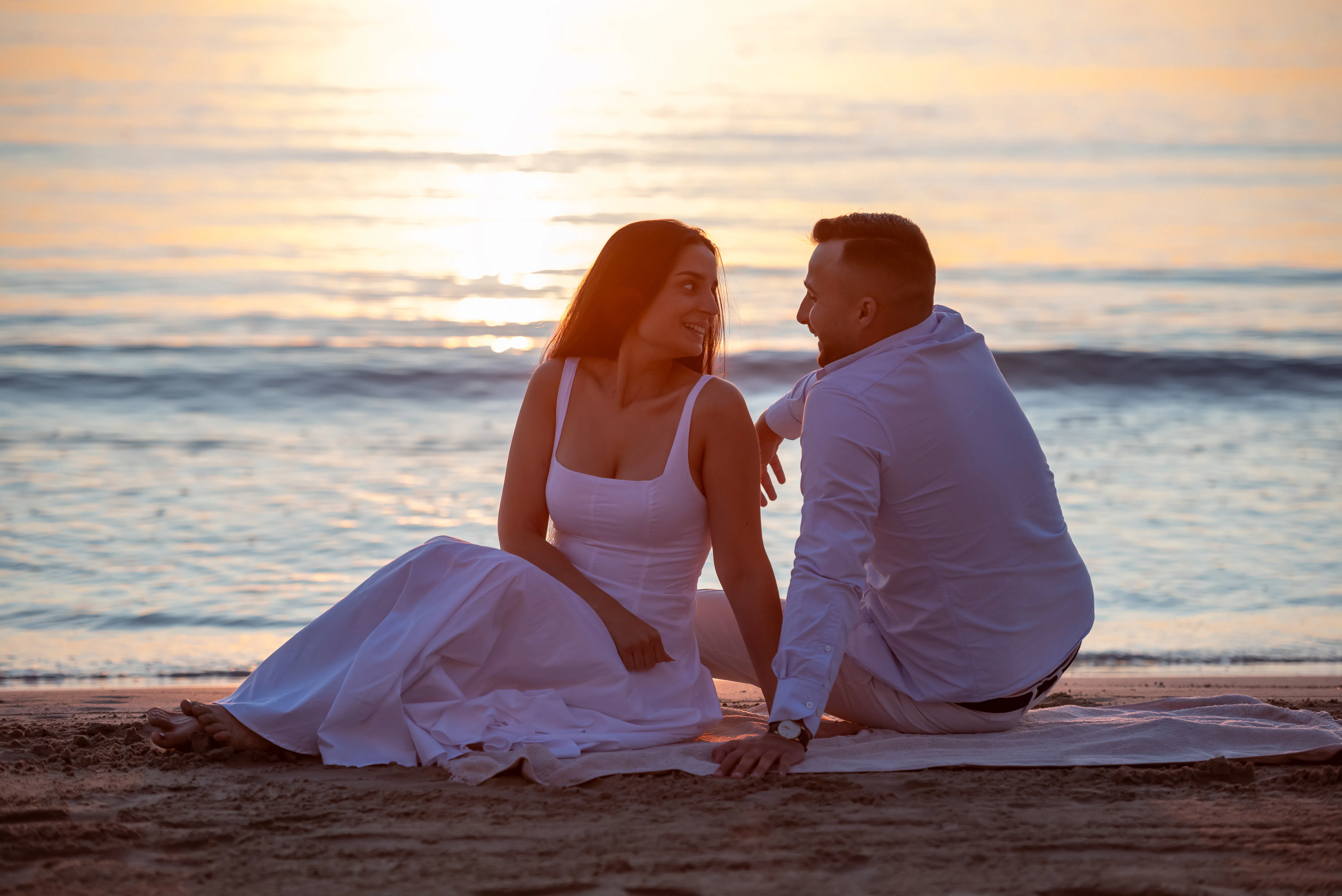 Young couple sitting on beach, looking at each other during golden hour at ocean