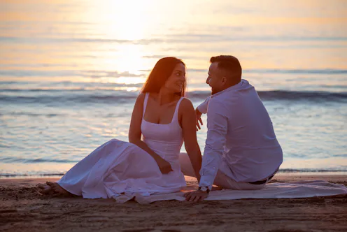 Young couple sitting on beach, looking at each other during golden hour at ocean