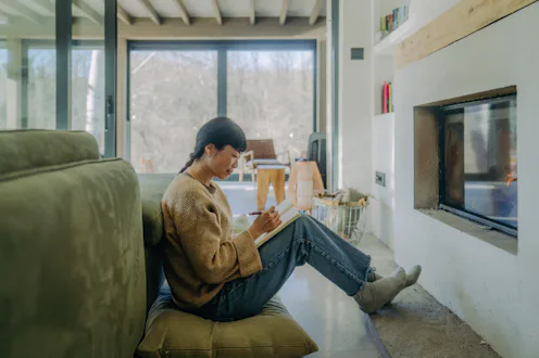 Photo of an Asian woman making an entry into her diary, while sitting by the cozy fireplace in her l...