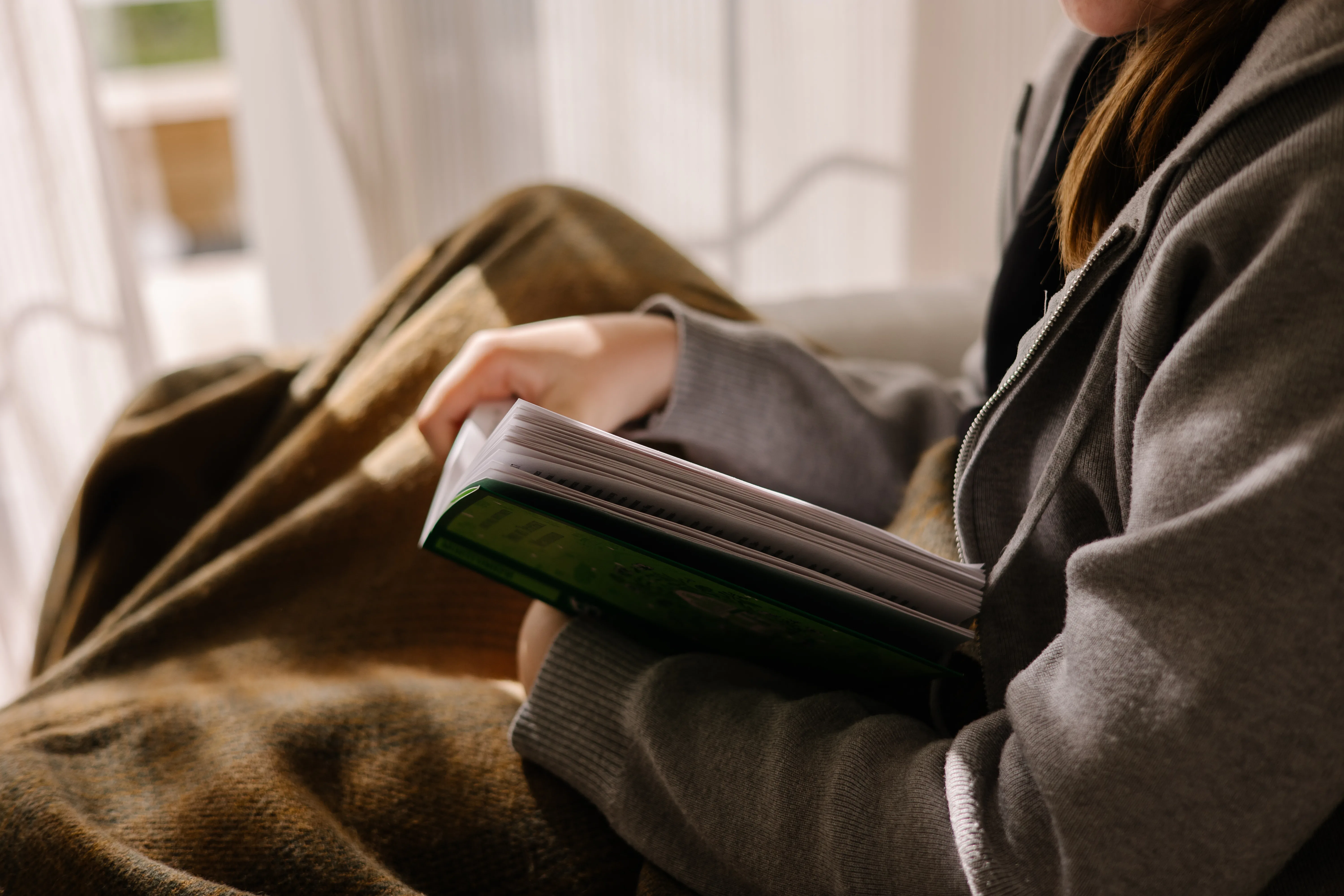 Teenage girl reading paper book while sitting wrapped in blanket near bright window in warm countrys...