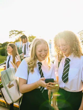 Teenage students in school uniforms sharing a laugh over a smartphone on stairs outdoors.