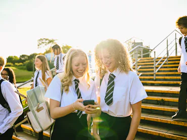 Teenage students in school uniforms sharing a laugh over a smartphone on stairs outdoors.