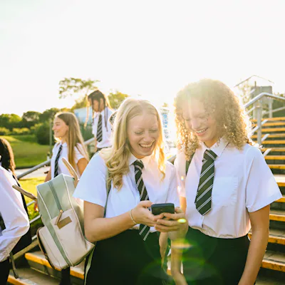 Teenage students in school uniforms sharing a laugh over a smartphone on stairs outdoors.