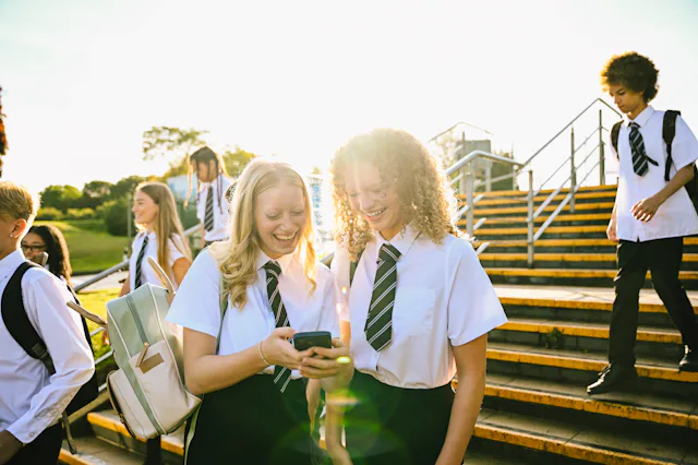 Teenage students in school uniforms sharing a laugh over a smartphone on stairs outdoors.