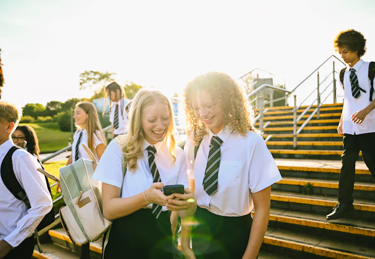 Teenage students in school uniforms sharing a laugh over a smartphone on stairs outdoors.