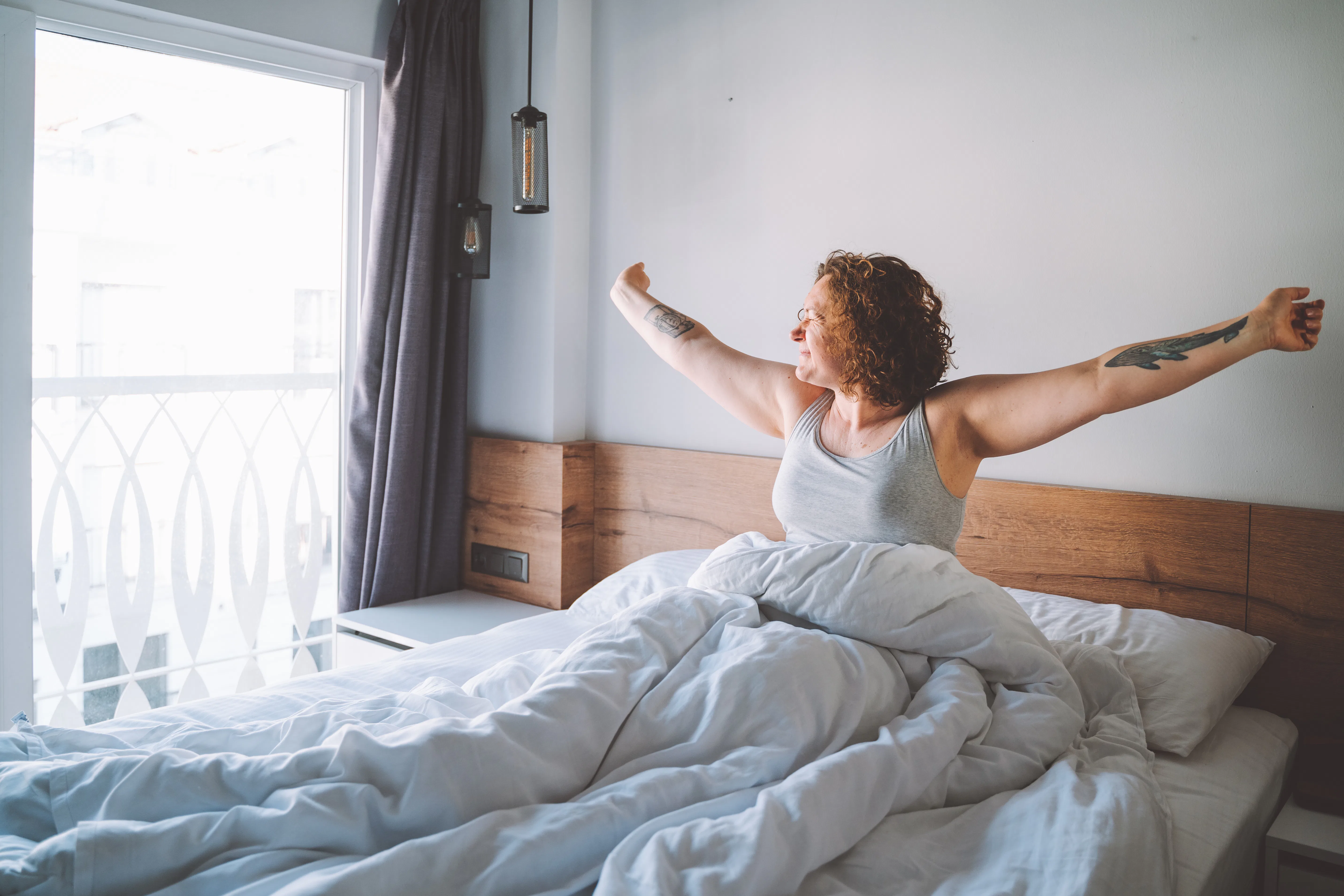 Middle age curly woman wake up in the morning full of happiness and strength and look to the window....