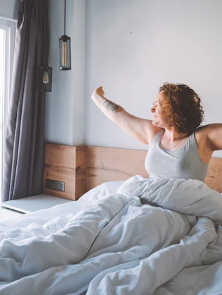 Middle age curly woman wake up in the morning full of happiness and strength and look to the window....