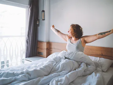 Middle age curly woman wake up in the morning full of happiness and strength and look to the window....