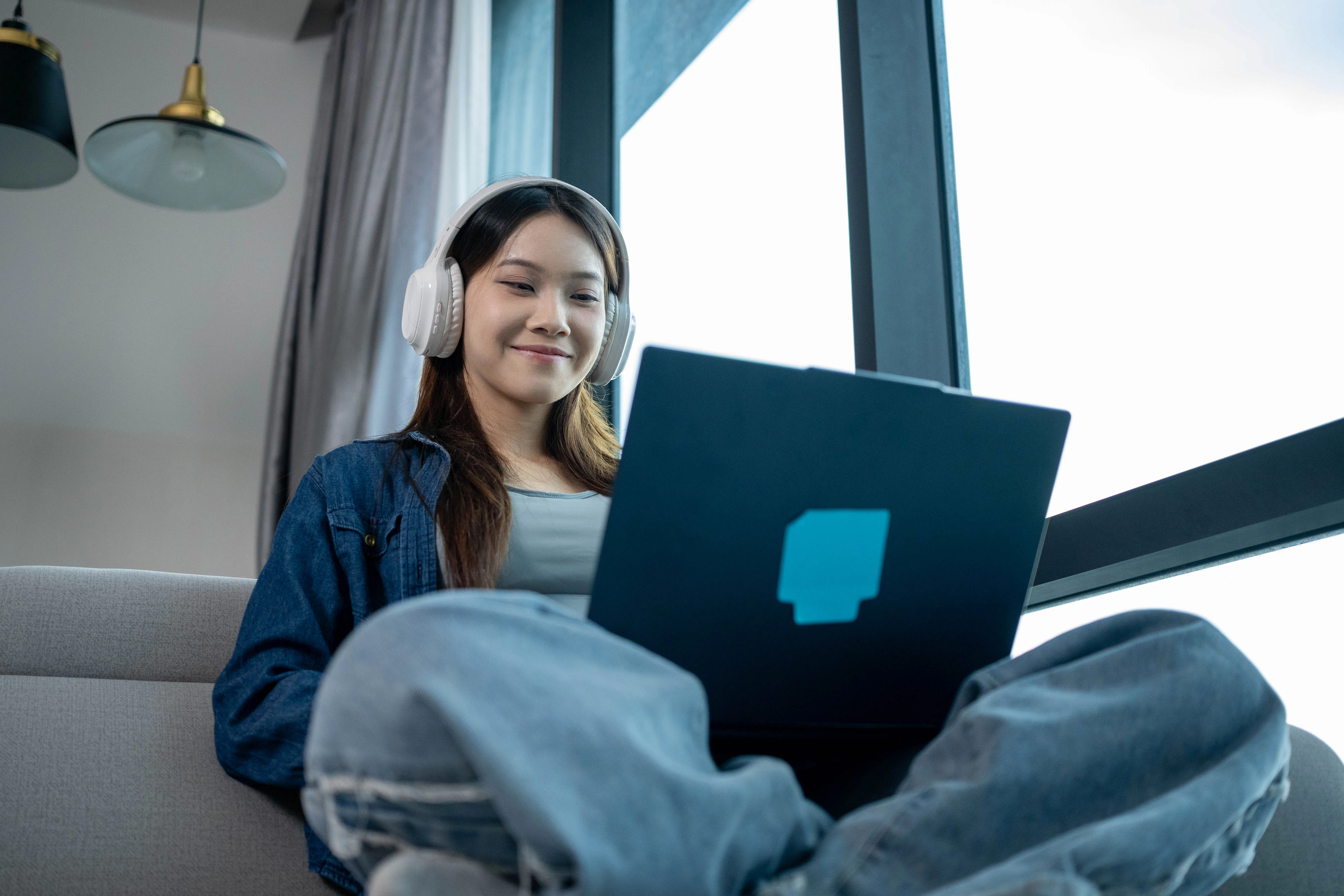 A cheerful Asian woman relaxes in a stylish living room while using her laptop computer, reflecting ...